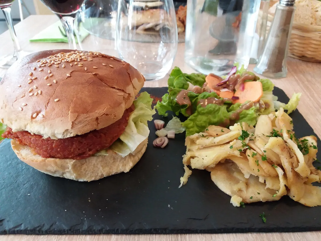 Un burger vegetariano con insalata e pastinaca su un tagliere di ardesia, con bicchieri e una saliera dietro