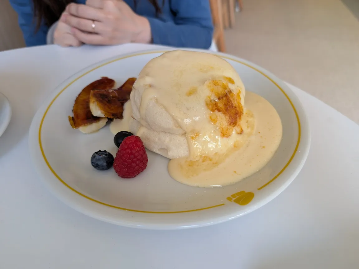 Una foto col telefono di pancake soufflé con crema pasticcera e frutti di bosco sul tavolo di un bar, con le mani di una persona dietro il piatto