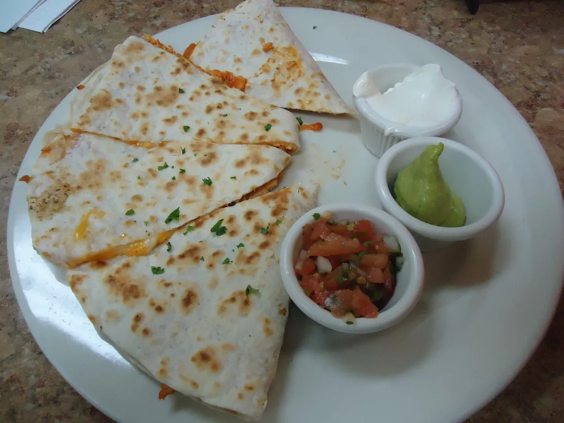 Una quesadilla di pollo con salsa, guacamole e panna acida su un piatto bianco, su un piano puntinato sotto una luce artificiale